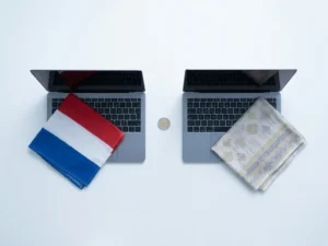 Two laptops on a white desk separated by a euro coin, one draped with a Dutch flag cloth, the other with Himalayan textile, flat lay view.