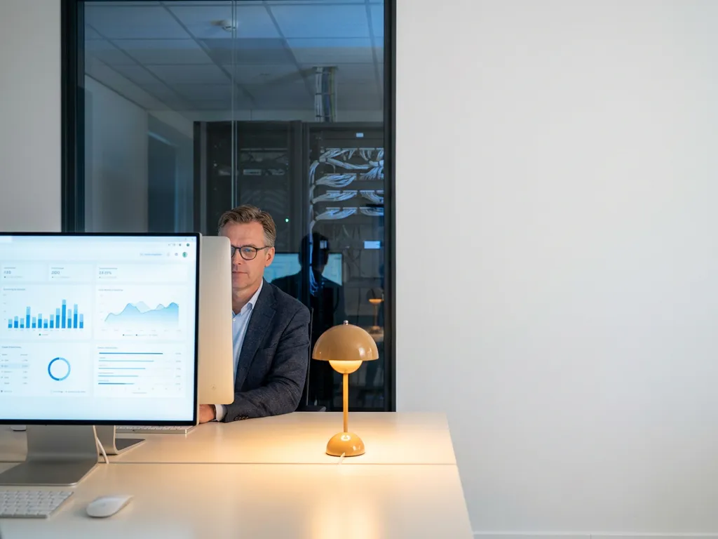 IT-manager bekijkt dashboardmetrics op groot scherm in modern kantoor met serverinfrastructuur op de achtergrond.