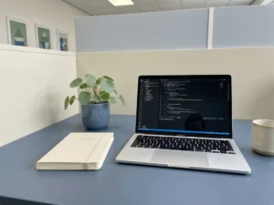 Bureau met laptop met schone code, notitieboek in het Nederlands en kleine kamerplant, symbool voor kennisbehoud binnen een organisatie.