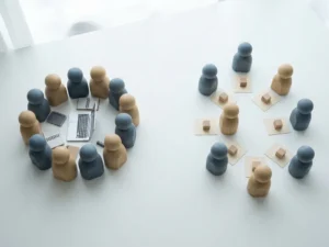 Miniature wooden figures split into two groups on a white desk, one clustered around a laptop, the other spread near individual task cards.