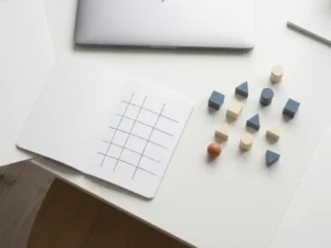 Overhead flat lay of a white desk with an open sprint-grid notebook, laptop, and geometric tokens arranged for distributed team planning.