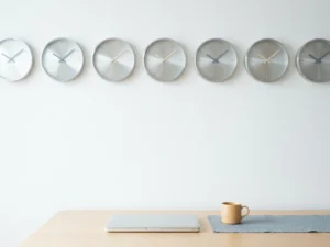Minimalist world clock display with multiple clock faces on a white wall showing different time zones above a modern desk with laptop and coffee cup.