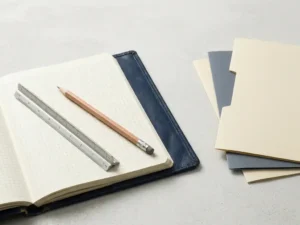 Open leren notitieboek met architectuurliniaal en potlood op betonnen bureau, omgeven door minimalistische documentmappen.