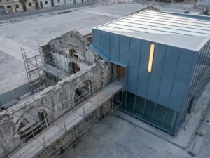 Verwerend stenen gebouw dat naadloos overgaat in een moderne glazen gevelstructuur, verbonden door steigers, fotografeerd vanuit een licht schuin bovenperspectief.