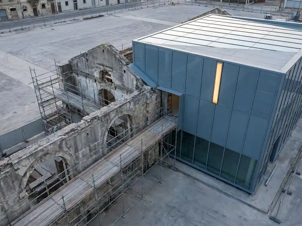Verwerend stenen gebouw dat naadloos overgaat in een moderne glazen gevelstructuur, verbonden door steigers, fotografeerd vanuit een licht schuin bovenperspectief.